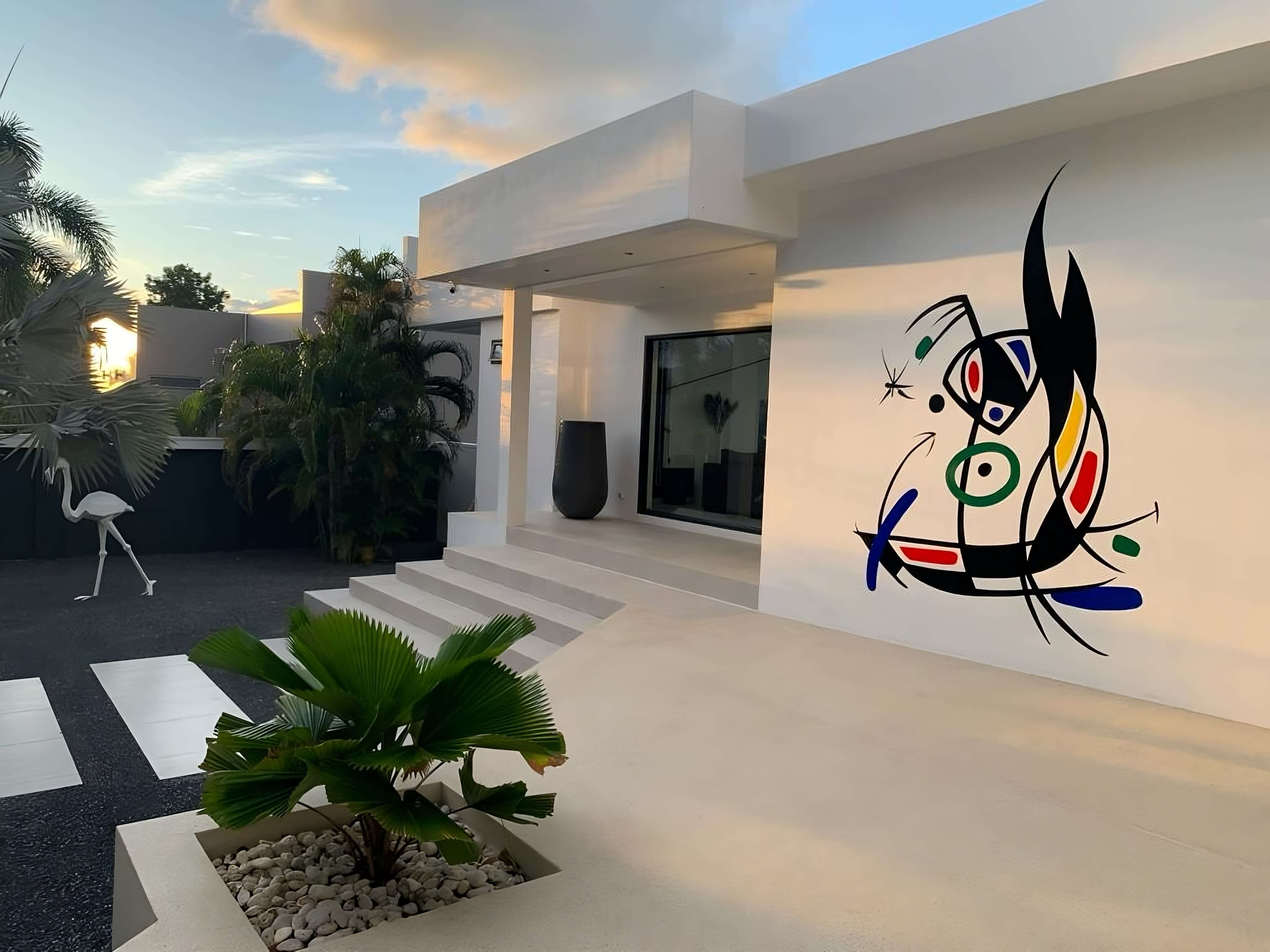 The villa entrance at golden hour — white architecture, palms, and a Miró-inspired mural beside the door.