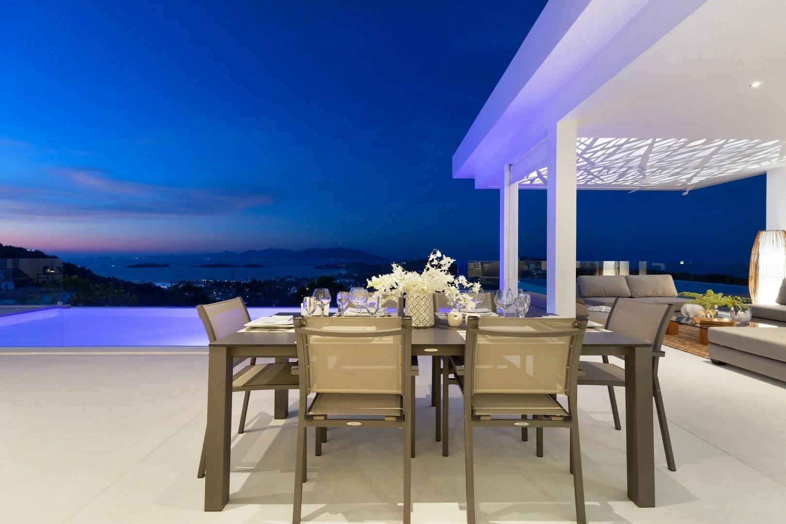 The summer dining table set with glasses and orchids at dusk, the bay fading to violet behind.