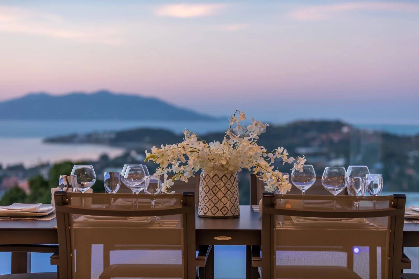 The dinner table set with wine glasses and orchids, dusk falling over the bay.